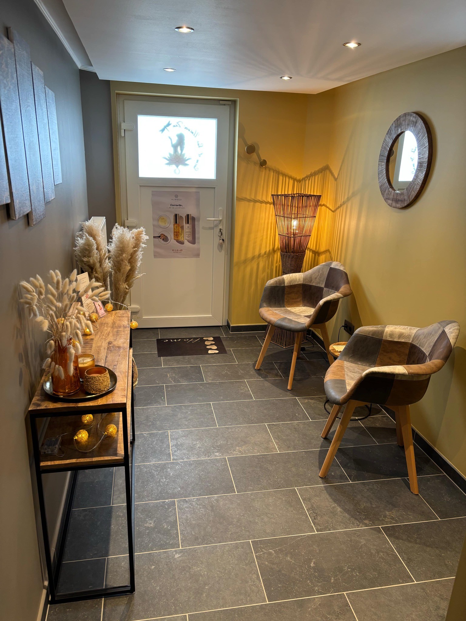 Cabine de soin avec table de massage drapée en rose pâle, serviettes enroulées décorées de petites roses, mur en lambris bois, plantes vertes suspendues et miroir organique doré